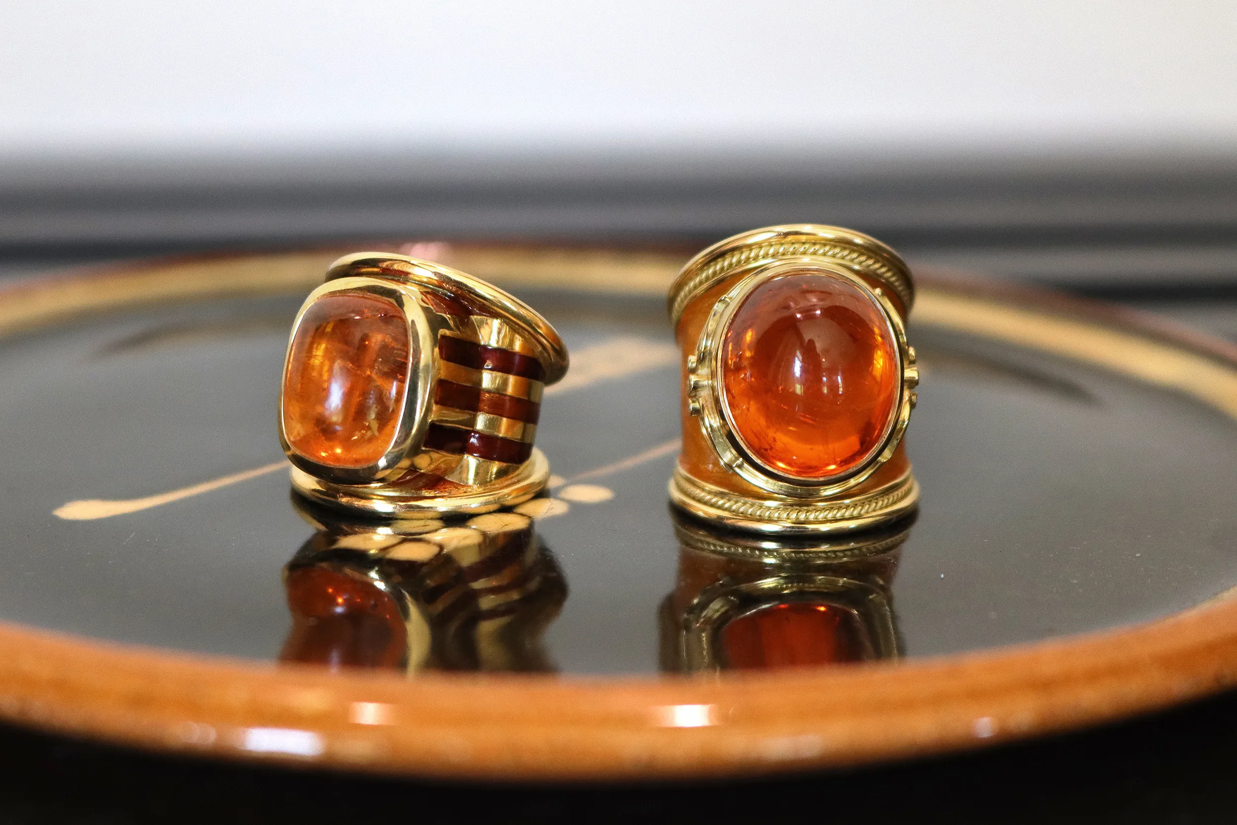 <p>Signature rings by Elizabeth Gage including the Aramis ring (left) with a 15.80 carat cushion-shaped sugarloaf mandarin garnet cabochon decorated with brown and orange enamel and the tapered Templar ring with a 29.37 carat oval-shaped mandarin garnet cabochon, brilliant-cut diamonds and saffron enamel, both in 18k yellow gold</p>