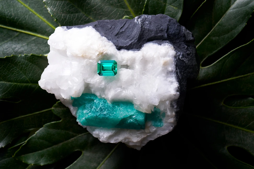 <p>An emerald-cut Muzo emerald sits on an emerald rough specimen, image courtesy of Thomas Levy</p>