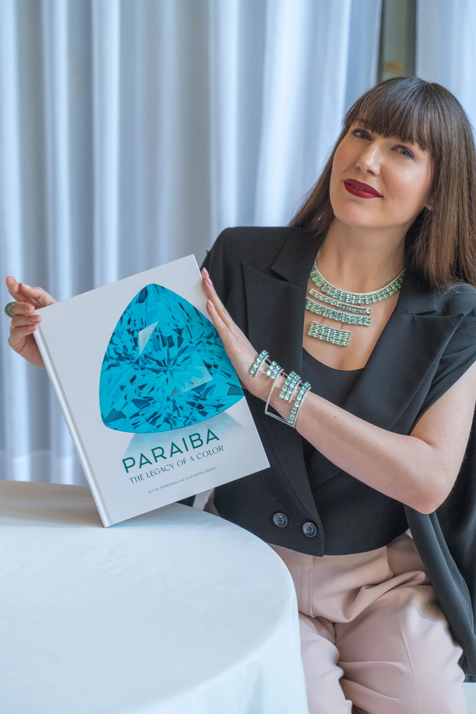 <p>Katerina Perez wearing a paraiba necklace and bracelet by Akillis, holding Paraiba – The Legacy of a Color</p>