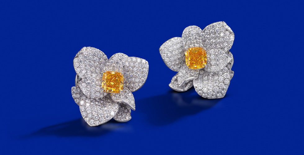 A rare pair of fancy coloured diamond and diamond earrings.  Each floral earring centring a radiant-cut fancy vivid yellowish orange diamond weighing 2.87 carats, and a radiant-cut fancy vivid yellow-orange diamond weighing 2.53 carats. Est. $ 360,00 – $ 490,000