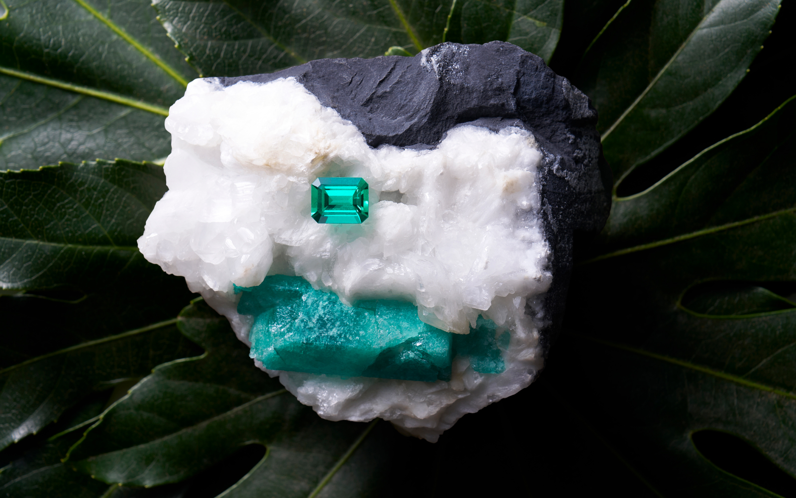An emerald-cut Muzo emerald sits on an emerald rough specimen, image courtesy of Thomas Levy