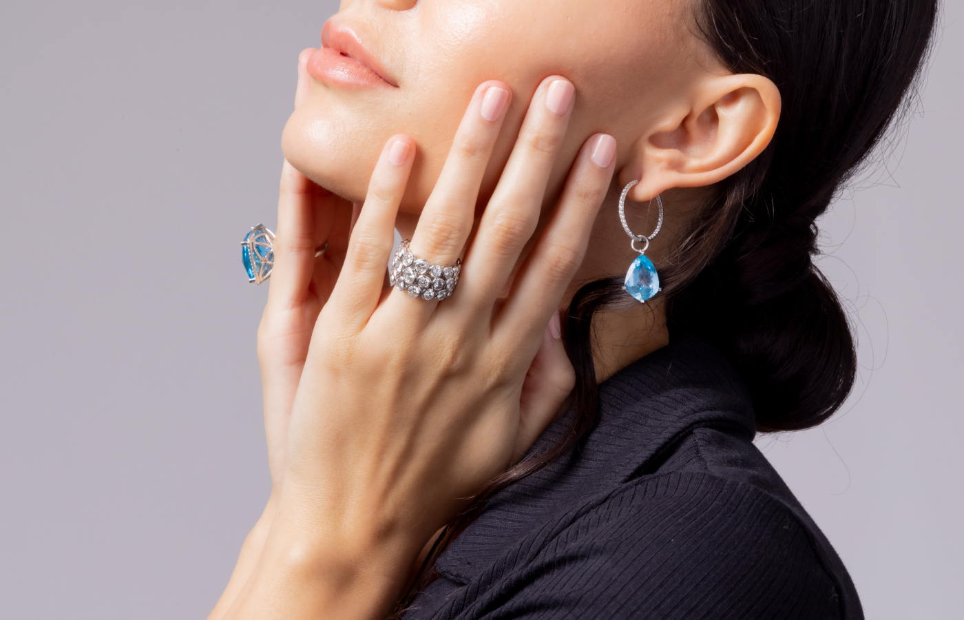 A model wears jewellery by Lisa Nik, including the Cushion ring from the Rocks collection with a blue topaz and diamonds in 18k white gold, a platinum ring from the Diamond Game collection with 7.82 carats of diamonds, and a pair of diamond hoop earrings with shield-shaped blue topaz charms from the Rocks and Sparkle collection  